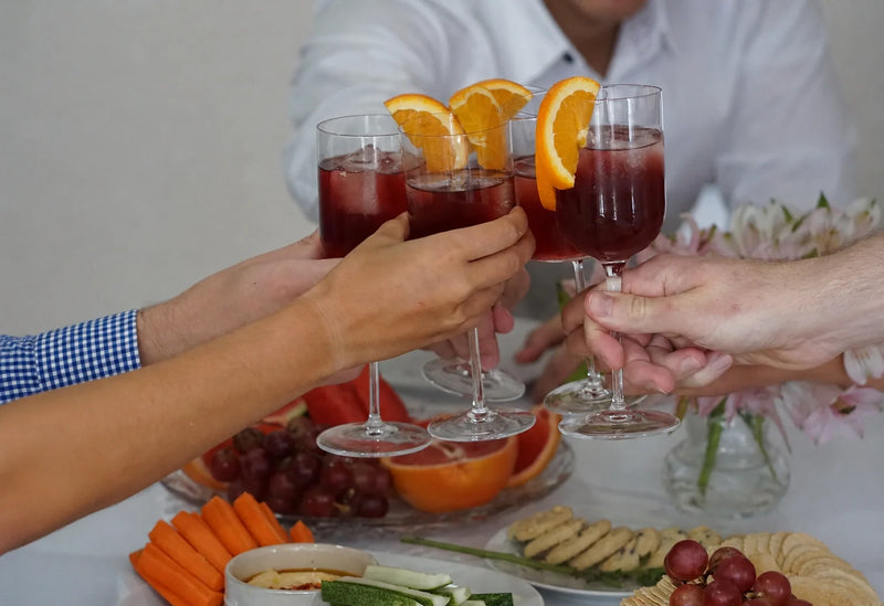 Friends toast with Melati botanical drinks and orange slices beside a colorful grazing table.