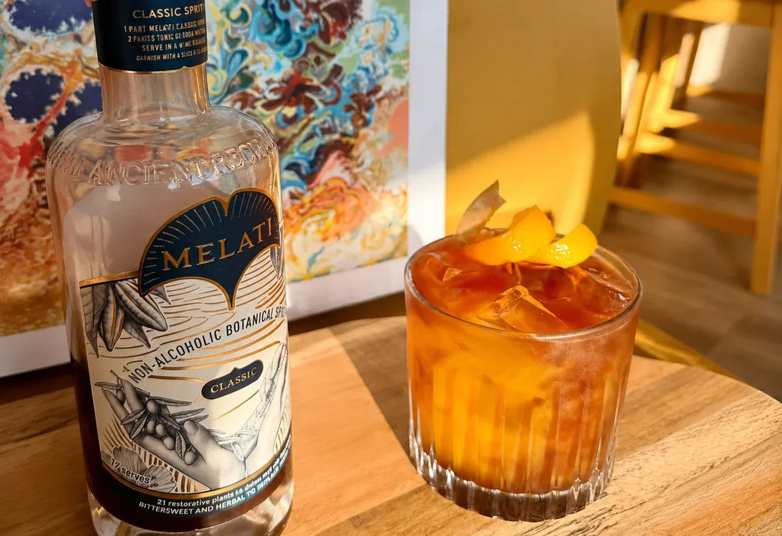 Melati Drinks Classic bottle beside an orange-garnished botanical drink on a sunlit wooden table.