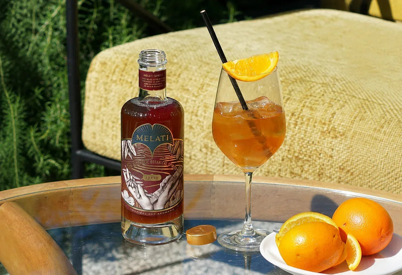 Melati Drinks Fresh bottle beside an orange-garnished botanical spritz on an outdoor table.